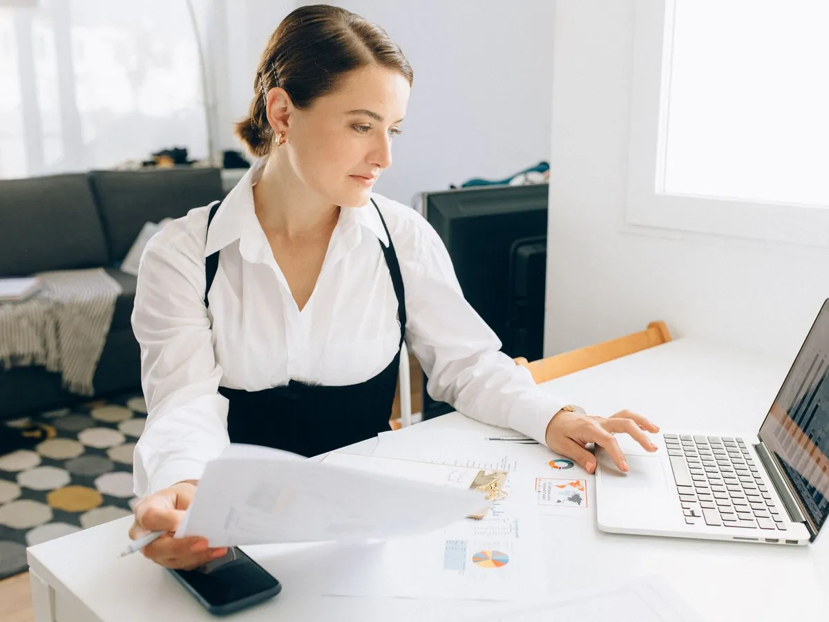 Empreendedora avaliando relatórios financeiros no notebook, com papéis e gráficos impressos na mesa