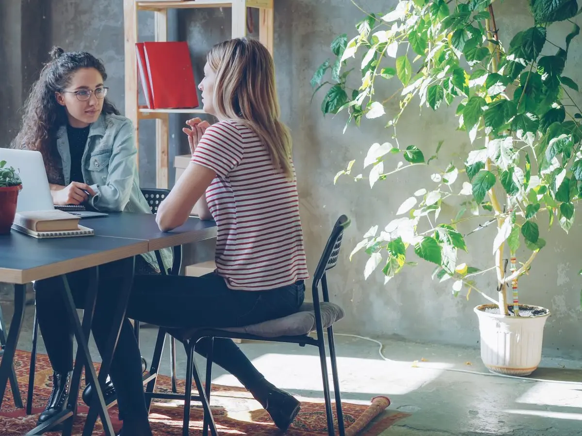 Consultora e cliente conversando em uma mesa de trabalho, com notebook e caderno de anotações abertos